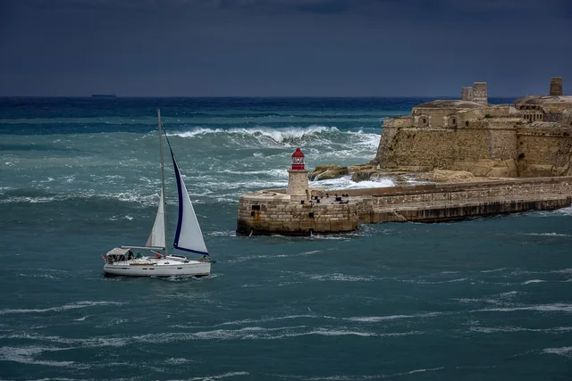 A yacht navigates the sea beside a stone breakwater and a red harbour light.