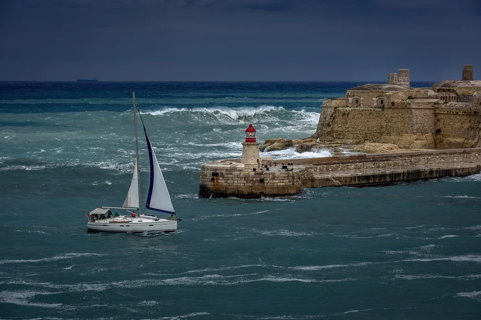 A yacht navigates the sea beside a stone breakwater and a red harbour light.